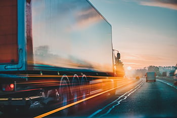 A truck driving into the sunset on a highway with reflections on the trailer.