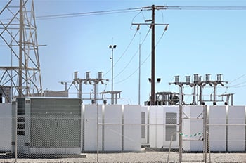 Battery storage solar farm, behind chain linked fence with electric transformers, against blue sky.