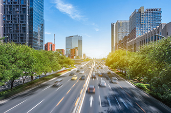 A cityscape featuring cars on a highway, trees, skyscrapers and blue sky