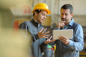 Two workers in blue clothing discussing information while looking at a white tablet