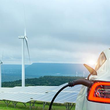 Electric vehicle charging in front of solar panels and wind turbines.