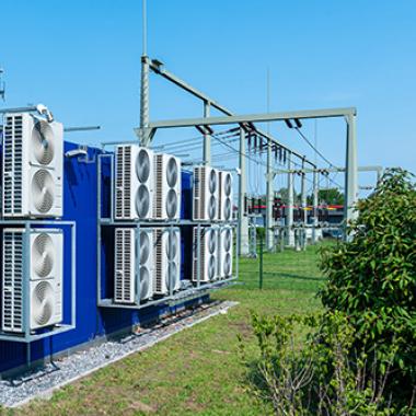 Transformer power station with large cooling fans on the rear wall against blue sky with green trees.