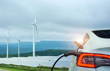 Electric vehicle charging in front of solar panels and wind turbines.