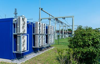 Transformer power station with large cooling fans on the rear wall against blue sky with green trees.
