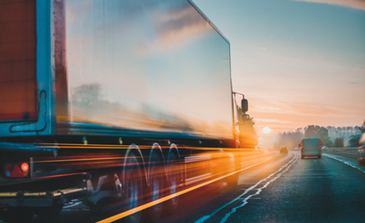 A truck driving into the sunset on a highway with reflections on the trailer.