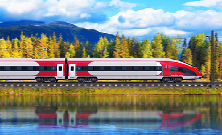 A white and red train driving from left to right at the border of a lake with trees and mountains in the background.
