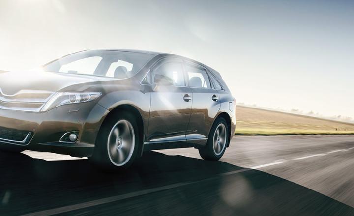 A grey car driving on the open road against a blue sky and sunshine.