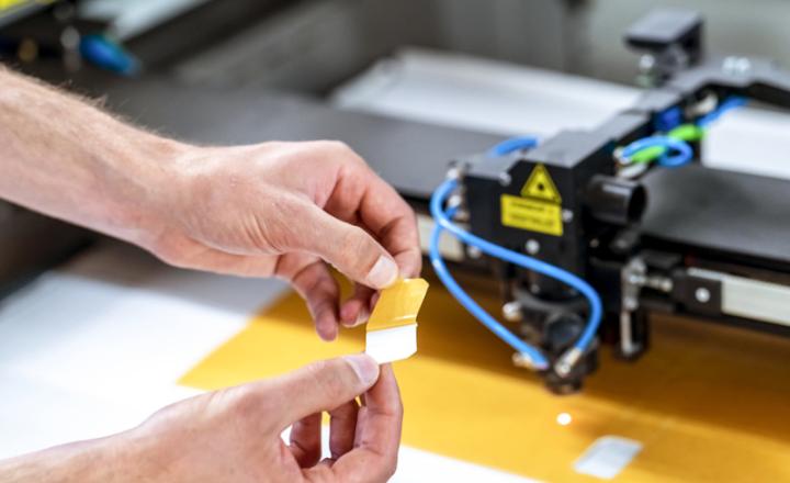 A pair of hands unpeeling yellow tape adhesive with machinery in the background