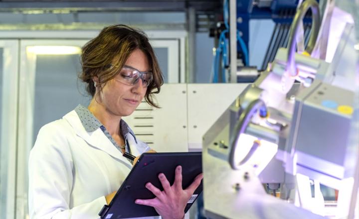 A technician wearing a white lab coat and safety glasses records data on a black tablet