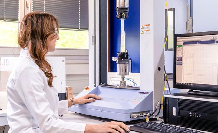 A woman in a white laboratory coat sits at a computer while conducting an experiment on a piece of adhesive tape