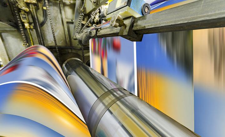 A large offset printing press running a long roll off paper over its rollers at high speed.
