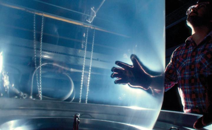 Man in dark manufacturing facility holding transparent, vertically blown film extrusion during heat-sealing process.