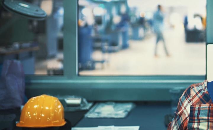 Production manager with a yellow hardhat in a factory control room looking through window onto production & manufacturing floor