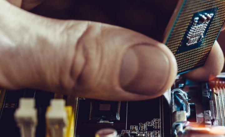 Hand holding circuitry chip over electronic computer motherboard with wires and nodes.