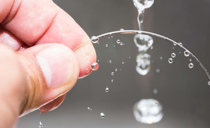 Hand holding a transparent, circular glass lens with clean water droplets streaming down its surface.