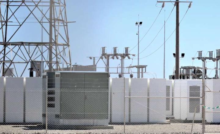 Battery storage solar farm, behind chain linked fence with electric transformers, against blue sky.