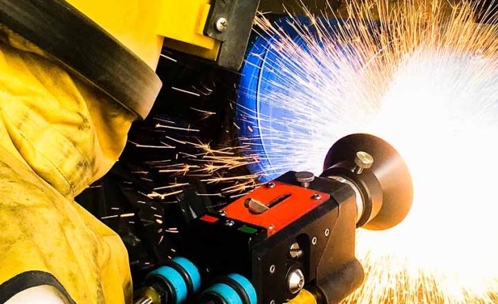 Technician wearing yellow protective gear and helmet with equipment during thermal spray process, with fiery white and orange sparks.