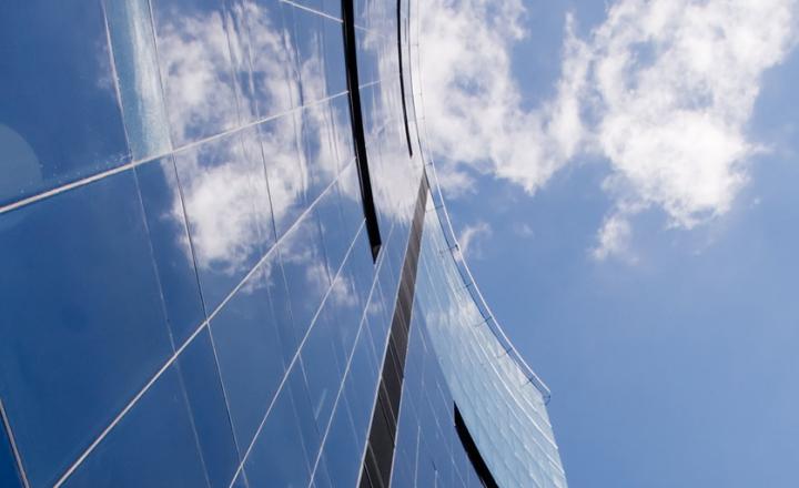 The side of a skyscraper building with panes of glass reflecting clouds and a blue sky.