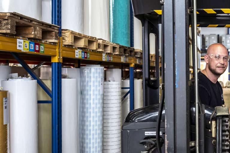 A forklift driver wearing safety glasses sits between two shelves of tape roll products on the warehouse production floor.