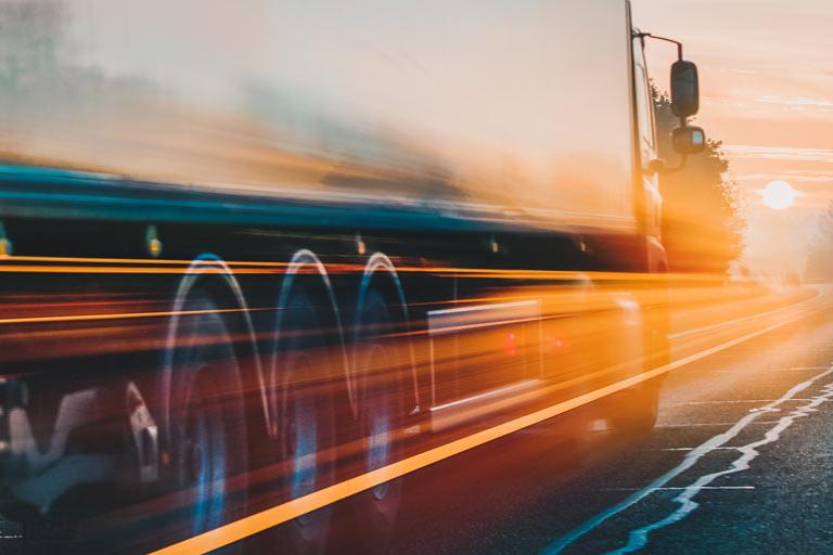 A truck driving into the sunset on a highway with reflections on the trailer.