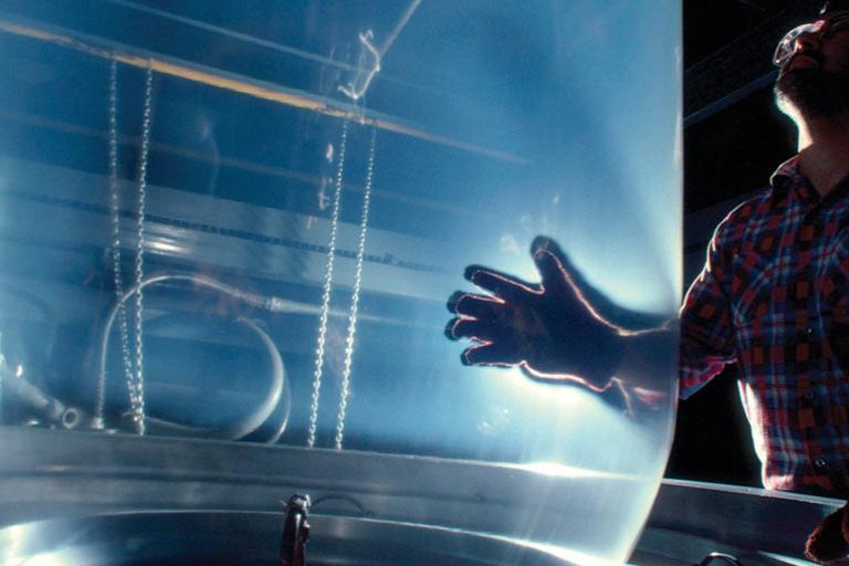 Man in dark manufacturing facility holding transparent, vertically blown film extrusion during heat-sealing process.