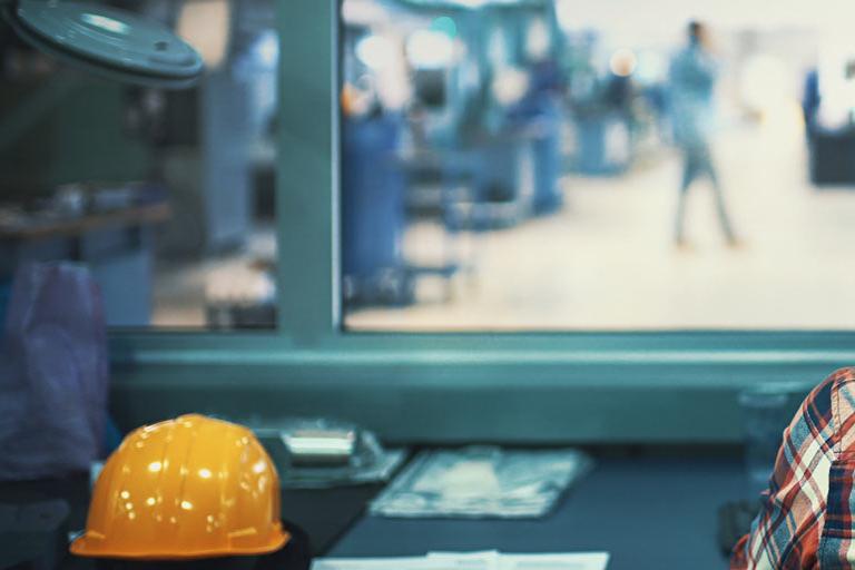 Production manager with a yellow hardhat in a factory control room looking through window onto production & manufacturing floor