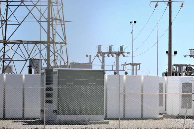 Battery storage solar farm, behind chain linked fence with electric transformers, against blue sky.