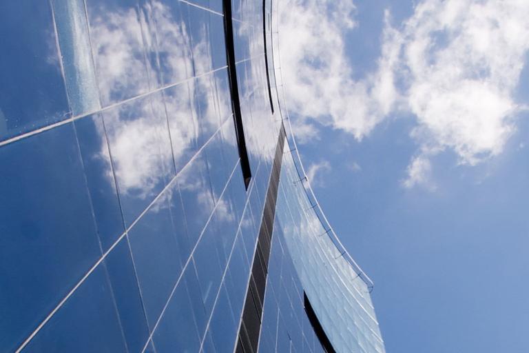 The side of a skyscraper building with panes of glass reflecting clouds and a blue sky.