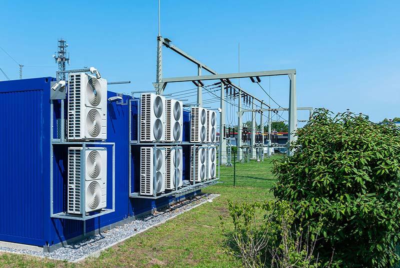 Transformer power station with large cooling fans on the rear wall against blue sky with green trees.